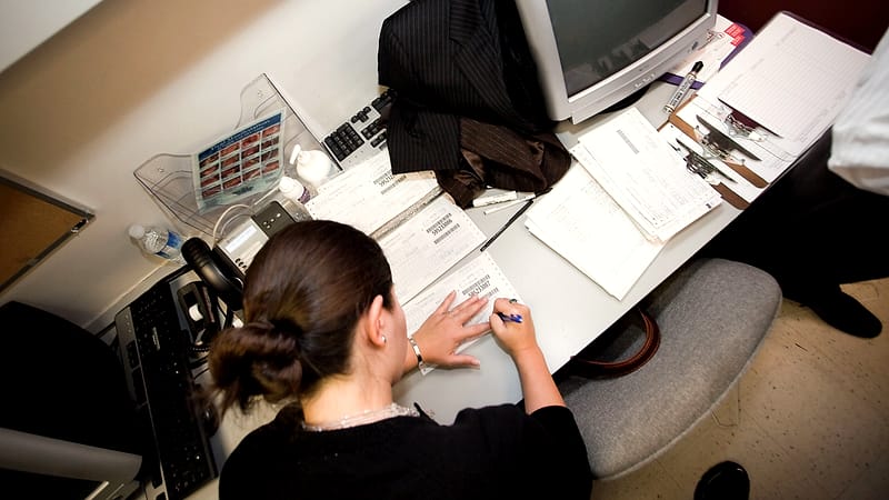 A nurse at a workstation entering information into a computer beside a stack of patient charts.