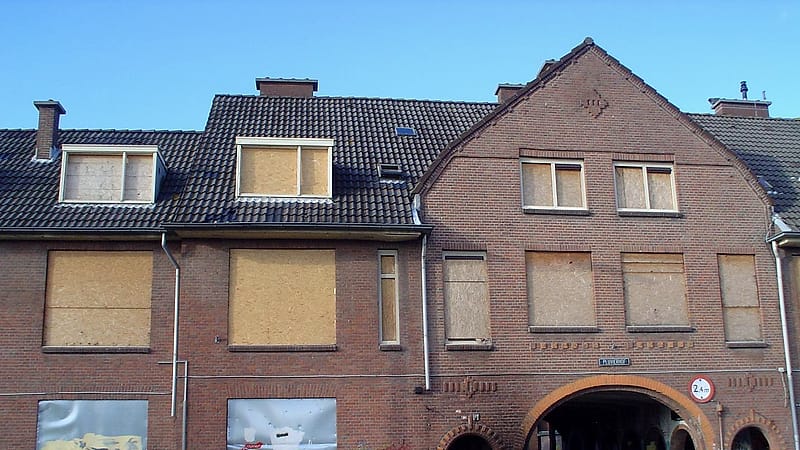 A boarded storefront with a collapsed section of cornice, water staining down the brick and weeds at the base.