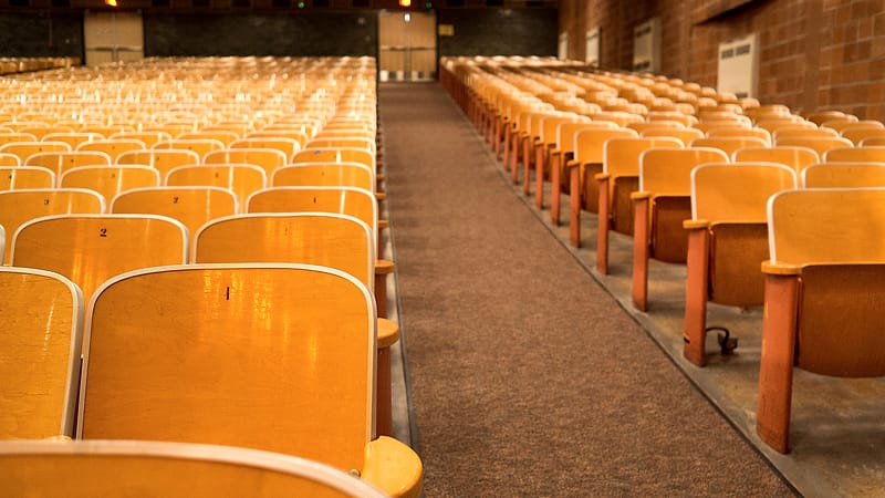 An empty wooden witness chair beside a microphone stand in a quiet paneled hearing room.