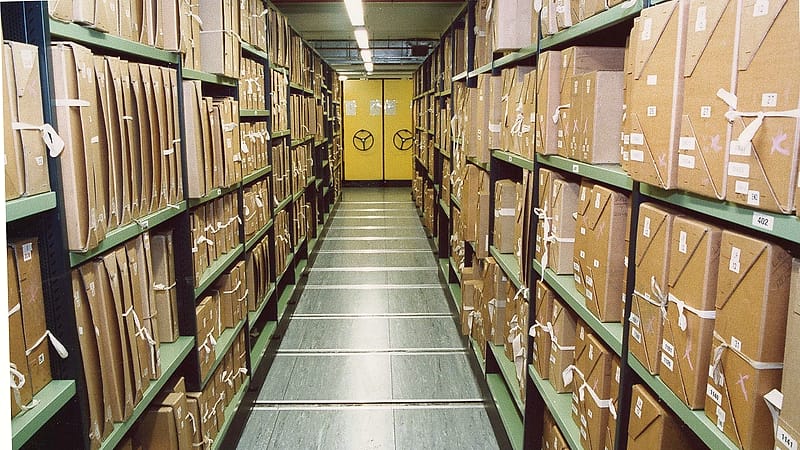 A shelved row of hardbound ledgers with handwritten spine labels in a storage room lit from one side.