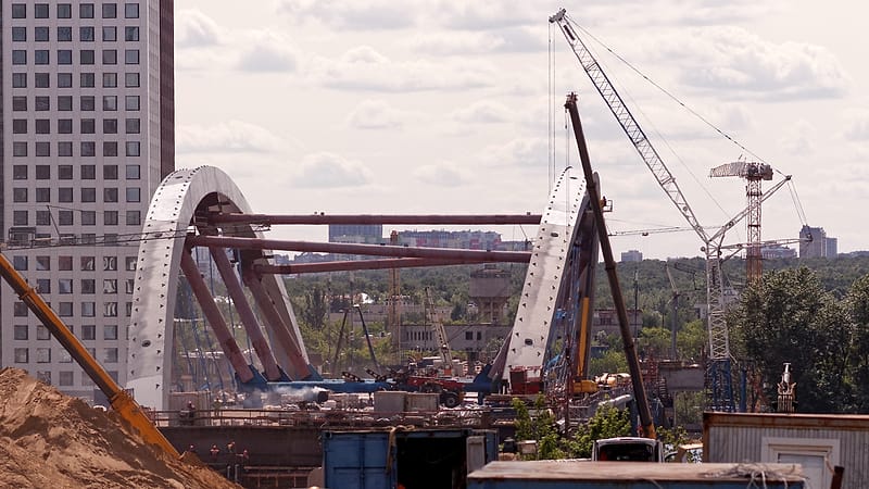 An excavator parked on bare graded soil beside orange safety fencing, with a row of older buildings behind it.