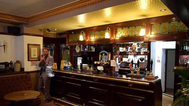 A narrow bar counter with stools on one side and a wall of shelved bottles behind it, warm light.