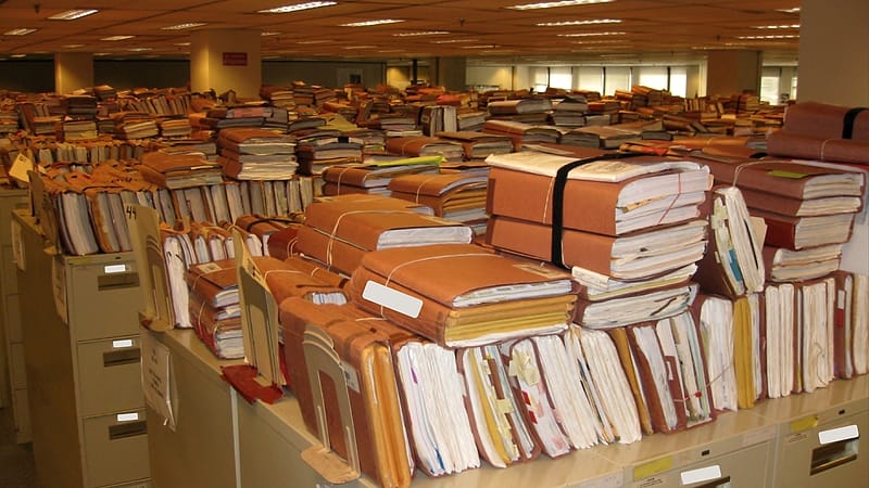 An open ring binder with tabbed dividers and a stack of stapled documents beside it on a wooden table.