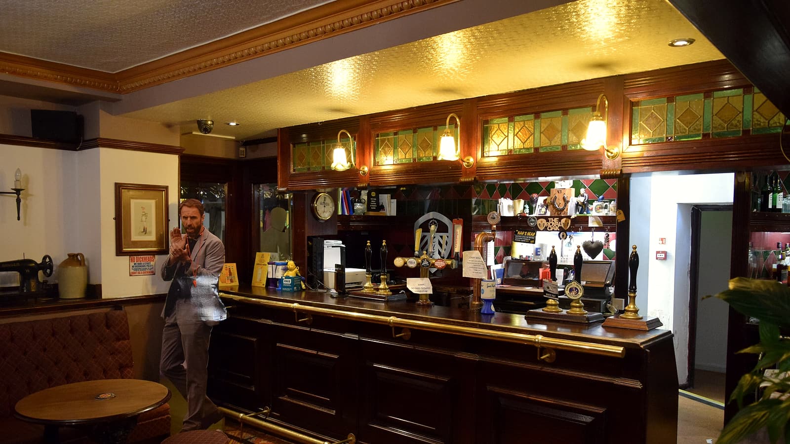 A narrow bar counter with stools on one side and a wall of shelved bottles behind it, warm light.
