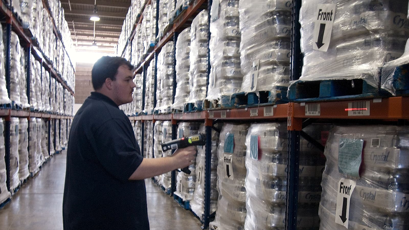 A handheld scanner reading a two-dimensional barcode on a small carton held over a conveyor.